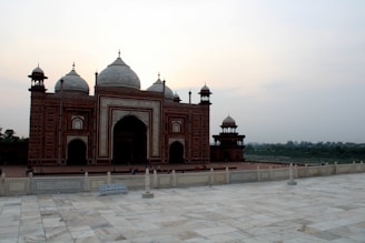 A beautifully detailed red sandstone building with intricate white marble inlays. The structure features several domes and towers, set against a backdrop of a gentle sky. The foreground has a marble platform with a sign and some tourists walking around.