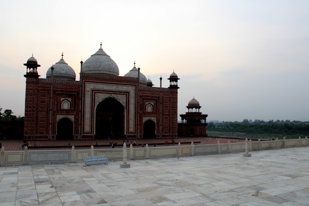 A beautifully detailed red sandstone building with intricate white marble inlays. The structure features several domes and towers, set against a backdrop of a gentle sky. The foreground has a marble platform with a sign and some tourists walking around.
