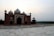 A beautifully detailed red sandstone building with intricate white marble inlays. The structure features several domes and towers, set against a backdrop of a gentle sky. The foreground has a marble platform with a sign and some tourists walking around.