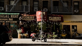 A street scene featuring several small shops with distinct signs, including a halal meat store, a florist with a red awning, and another shop in yellow. In front of the florist, there are various flowers and plants displayed for sale. Two scooters are parked on the sidewalk, with a bike in the foreground. Shadows suggest late afternoon lighting.