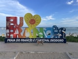 A colorful sign with large, artistic letters spelling out 'Eu Amo Francês' is situated on a sandy beach. Each letter is decorated with various symbols such as a sailboat, palm tree, and sun. In the background, people are enjoying the beach and the ocean under a partly cloudy blue sky. A dog is standing on the sand in front of the sign.