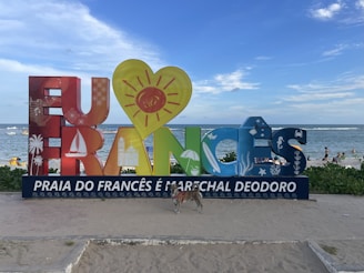A colorful sign with large, artistic letters spelling out 'Eu Amo Francês' is situated on a sandy beach. Each letter is decorated with various symbols such as a sailboat, palm tree, and sun. In the background, people are enjoying the beach and the ocean under a partly cloudy blue sky. A dog is standing on the sand in front of the sign.