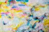 A textured paper surface is covered with colorful abstract watercolor patterns. Vivid splashes of yellow, blue, pink, and black blend and bleed into each other, creating a dynamic and artistic look.
