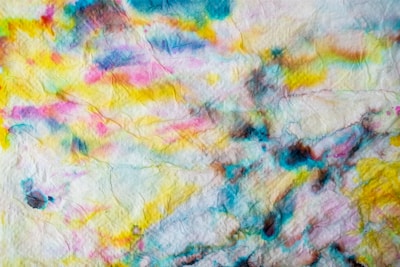 A textured paper surface is covered with colorful abstract watercolor patterns. Vivid splashes of yellow, blue, pink, and black blend and bleed into each other, creating a dynamic and artistic look.