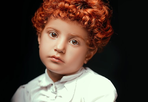A young child with curly red hair and expressive eyes gazes directly at the camera. The child is wearing a white collared shirt, set against a dark background, which enhances the focus on their facial expression.