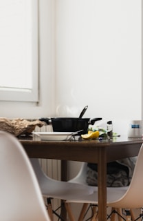 A wooden kitchen table is set with a steaming black pot and a lid resting on top. Beside it, there are white chairs and miscellaneous kitchen items, including a cut lemon, a white bowl with cutlery, and a crumpled paper bag. The scene is bathed in natural light from a nearby window.