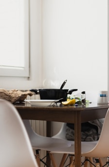 A wooden kitchen table is set with a steaming black pot and a lid resting on top. Beside it, there are white chairs and miscellaneous kitchen items, including a cut lemon, a white bowl with cutlery, and a crumpled paper bag. The scene is bathed in natural light from a nearby window.