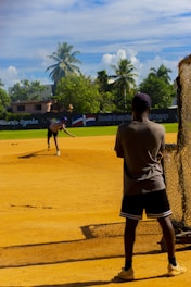Two people are on a baseball field, one pitching and the other standing near a batting net. The field is surrounded by greenery including palm trees and there are buildings in the background. The sky is bright with blue color and scattered clouds.