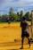 Two people are on a baseball field, one pitching and the other standing near a batting net. The field is surrounded by greenery including palm trees and there are buildings in the background. The sky is bright with blue color and scattered clouds.