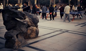 People are playing table tennis in an open area, surrounded by tall trees. There is a large rock formation in the foreground, and several individuals can be seen holding paddles and watching the game.
