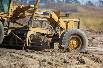 A large yellow construction grader is working on a dirt road, leveling the ground. The scene shows dirt being moved by the blade of the machine, with a clear blue sky and blurred outlines of trees in the background.