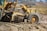 A large yellow construction grader is working on a dirt road, leveling the ground. The scene shows dirt being moved by the blade of the machine, with a clear blue sky and blurred outlines of trees in the background.