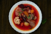 A bowl filled with a vibrant red soup containing pieces of beetroot, carrots, meat, cauliflower, and potatoes. The soup is garnished with fresh herbs and presented in a white dish.