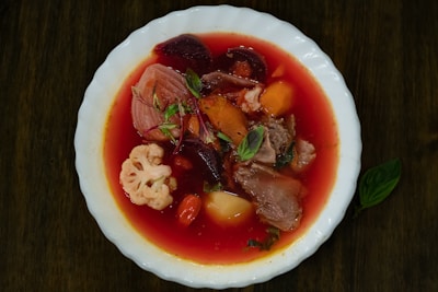 A bowl filled with a vibrant red soup containing pieces of beetroot, carrots, meat, cauliflower, and potatoes. The soup is garnished with fresh herbs and presented in a white dish.