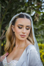 A bride with long blonde hair is wearing a white wedding dress and a sheer veil adorned with decorative elements. She has her eyes closed and her makeup is elegantly done with bold eyebrows, eye shadow, and lipstick. She is standing outdoors surrounded by greenery.