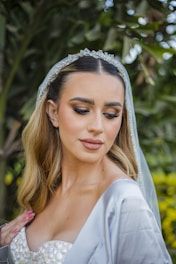 A bride with long blonde hair is wearing a white wedding dress and a sheer veil adorned with decorative elements. She has her eyes closed and her makeup is elegantly done with bold eyebrows, eye shadow, and lipstick. She is standing outdoors surrounded by greenery.