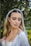 A bride with long blonde hair is wearing a white wedding dress and a sheer veil adorned with decorative elements. She has her eyes closed and her makeup is elegantly done with bold eyebrows, eye shadow, and lipstick. She is standing outdoors surrounded by greenery.