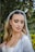 A bride with long blonde hair is wearing a white wedding dress and a sheer veil adorned with decorative elements. She has her eyes closed and her makeup is elegantly done with bold eyebrows, eye shadow, and lipstick. She is standing outdoors surrounded by greenery.