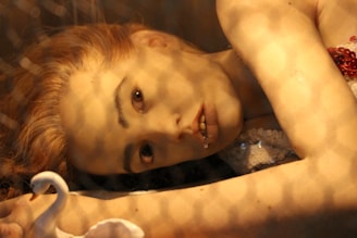 A person lies down with an intense gaze, surrounded by a net-like shadow pattern. The face is close to the camera, displaying an expression that blends curiosity and enigma. Next to them is a white swan figure, adding an artistic and surreal touch. The lighting casts a warm, amber glow over the scene.