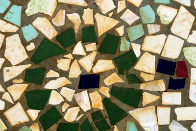 A mosaic pattern composed of irregularly shaped tiles in various colors and sizes. The tiles are primarily shades of beige, green, blue, and dark blue, creating a fragmented yet visually harmonious design.