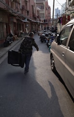 A narrow urban street bordered by older brick buildings, with laundry hanging between them. A person in a camouflage jacket walks away, dragging a suitcase. Several scooters are parked alongside the street, and some vehicles, including a white van, are visible. The early evening sunlight casts long shadows.