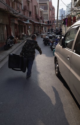 A narrow urban street bordered by older brick buildings, with laundry hanging between them. A person in a camouflage jacket walks away, dragging a suitcase. Several scooters are parked alongside the street, and some vehicles, including a white van, are visible. The early evening sunlight casts long shadows.