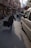 A narrow urban street bordered by older brick buildings, with laundry hanging between them. A person in a camouflage jacket walks away, dragging a suitcase. Several scooters are parked alongside the street, and some vehicles, including a white van, are visible. The early evening sunlight casts long shadows.