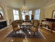 A luxurious parlor with ornate furniture, including five upholstered chairs arranged around a small table with a tea set. An elaborate chandelier hangs from the ceiling. The room features large windows with white drapes, ornate moldings, a grand fireplace on the left, and a grand piano on the right.