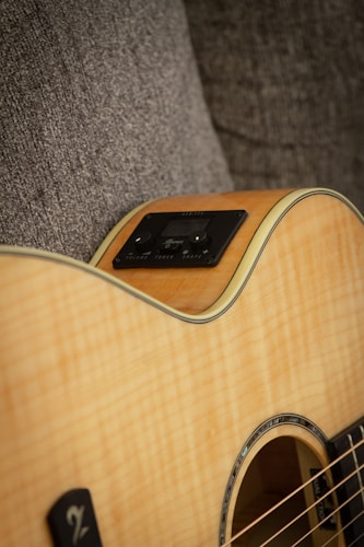 A close-up view of an acoustic guitar highlighting its natural wood finish and intricate details such as the control panel and soundhole. The guitar is placed on a textured gray fabric background.