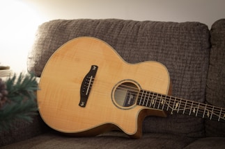 An acoustic guitar with a natural wood finish is resting on a textured, grey-brown couch. The lighting is soft, creating a warm and cozy atmosphere. A part of a green plant is visible in the lower left corner, and the background wall is clean and minimalistic.