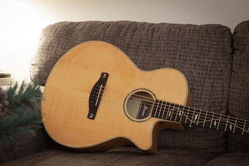 An acoustic guitar with a natural wood finish is resting on a textured, grey-brown couch. The lighting is soft, creating a warm and cozy atmosphere. A part of a green plant is visible in the lower left corner, and the background wall is clean and minimalistic.