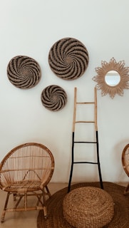 A cozy interior setting featuring wicker furniture, including two chairs, a circular ottoman, and a wall-mounted decor piece composed of three round woven baskets and a sun-shaped mirror. A wooden ladder with black accents leans against the wall.
