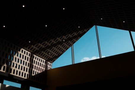 The interior features a modern architectural design with a high, angular ceiling made from dark materials. Large windows allow natural light to illuminate the space, revealing a blue sky and a few clouds outside. The light creates interesting geometric shadows and patterns on the walls.