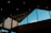 The interior features a modern architectural design with a high, angular ceiling made from dark materials. Large windows allow natural light to illuminate the space, revealing a blue sky and a few clouds outside. The light creates interesting geometric shadows and patterns on the walls.