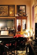 A vintage-style room filled with antique furniture and decorations. The center features a wooden piano with sheet music on its stand, beside a chair with a red cloth. Above, various paintings and artwork are displayed, adding an artistic ambiance. A mirror reflects part of the room and a person in a red garment standing near the doorway. Soft lighting casts a warm glow over the scene, creating an intimate atmosphere.