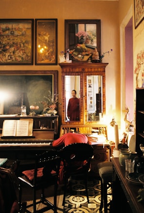 A vintage-style room filled with antique furniture and decorations. The center features a wooden piano with sheet music on its stand, beside a chair with a red cloth. Above, various paintings and artwork are displayed, adding an artistic ambiance. A mirror reflects part of the room and a person in a red garment standing near the doorway. Soft lighting casts a warm glow over the scene, creating an intimate atmosphere.