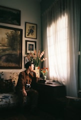 A person sits on a sofa in a cozy, art-filled room, gazing out of a window. The room is decorated with framed artwork on the walls and a vase of flowers on a table. Natural light streams through sheer curtains, creating a warm and contemplative atmosphere.