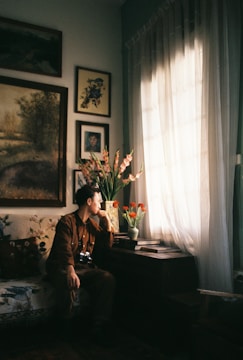 A person sits on a sofa in a cozy, art-filled room, gazing out of a window. The room is decorated with framed artwork on the walls and a vase of flowers on a table. Natural light streams through sheer curtains, creating a warm and contemplative atmosphere.
