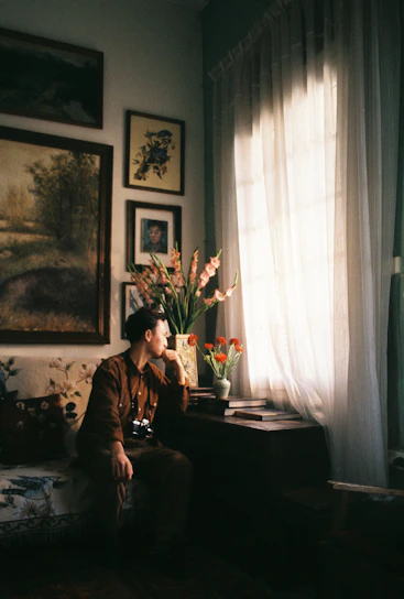 A warm portrait of a thoughtful woman in a cozy, sunlit room filled with plants and architectural sketches.