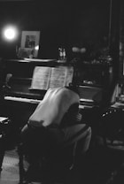 A dimly lit room with a grand piano topped with sheet music, partially illuminated by a bright light source. A shirtless person with their back to the camera is sitting on a bench, appearing to lean forward with their arms around their knees. The ambiance is moody and introspective, enhanced by the shadows and subdued lighting.
