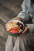 A person wearing a denim jacket and grey pants holds a bowl containing a colorful salad. The salad includes quinoa, chickpeas, roasted tomatoes, nuts, and dark cubes of what appears to be beets.