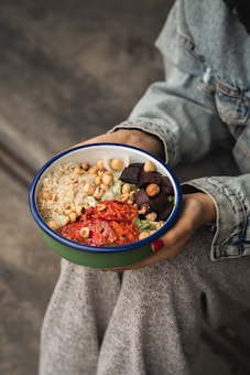 A person wearing a denim jacket and grey pants holds a bowl containing a colorful salad. The salad includes quinoa, chickpeas, roasted tomatoes, nuts, and dark cubes of what appears to be beets.