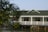 A house with a green roof and siding is situated partially over a body of water. The structure features a white railing along a porch or deck area. Trees with green foliage frame the foreground of the scene.
