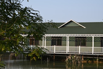 A house with a green roof and siding is situated partially over a body of water. The structure features a white railing along a porch or deck area. Trees with green foliage frame the foreground of the scene.