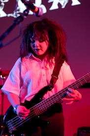 A musician with dramatic makeup and voluminous hair plays an electric bass guitar. The setting is dimly lit with a predominantly red and purple backdrop. The musician is wearing a white shirt, and there is a focused expression while performing. A microphone and amplifier are also visible in the background.