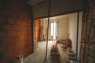 An unfinished room with exposed brickwork on the left, partially plastered walls, and construction materials scattered across the floor. A large window at the far end provides natural light. Tools and buckets are present, suggesting ongoing renovation work.