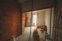 An unfinished room with exposed brickwork on the left, partially plastered walls, and construction materials scattered across the floor. A large window at the far end provides natural light. Tools and buckets are present, suggesting ongoing renovation work.