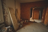 The image depicts an unfinished interior space with exposed brick walls and a concrete floor. Construction materials such as wooden boards, buckets, and other building supplies are scattered around the room. There is a partially opened doorway leading to another room, highlighting the ongoing renovation process.