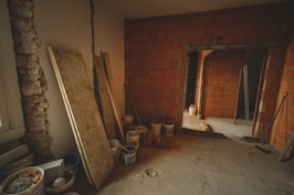 The image depicts an unfinished interior space with exposed brick walls and a concrete floor. Construction materials such as wooden boards, buckets, and other building supplies are scattered around the room. There is a partially opened doorway leading to another room, highlighting the ongoing renovation process.