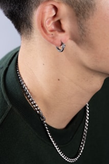 A close-up of a person's neck and ear, showing a silver hoop earring in the ear and a silver chain necklace around the neck. The person is wearing a green garment.